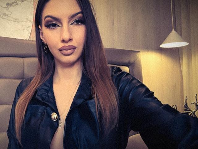 LucyGrace with long brown hair, wearing a black satin jacket, has pouty lips and dark eyeliner, showing her tits slightly in a low-cut top. Her ass is not visible.