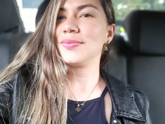 MelParker has long, straight brown hair, pink lipstick, and wears a black leather jacket over a black top with a gold necklace. She's smiling slightly with her head tilted.