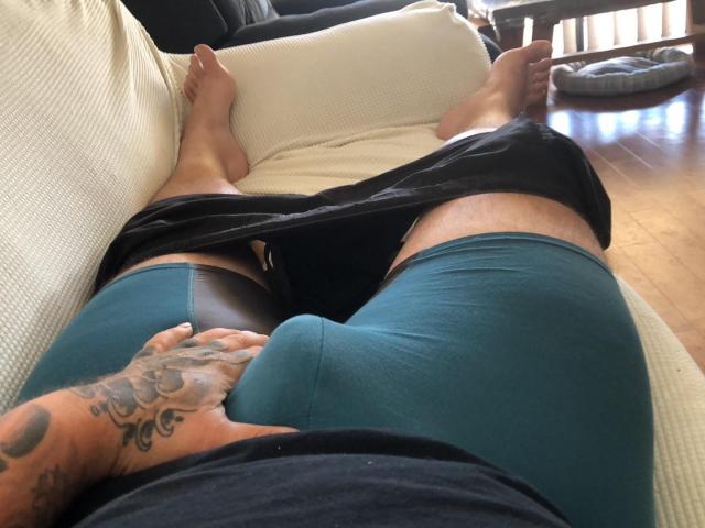 AussieStrong's legs, wearing teal shorts, black underwear pulled down, tattooed hand on crotch, feet on white pillow, wooden floor, black shirt, daytime, relaxed, casual.