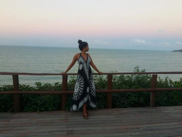 LunaMelynda-hot stands on a wooden deck, hands on a rail, wearing a black and white patterned halter dress, with her hair in a bun, looking at the ocean.
