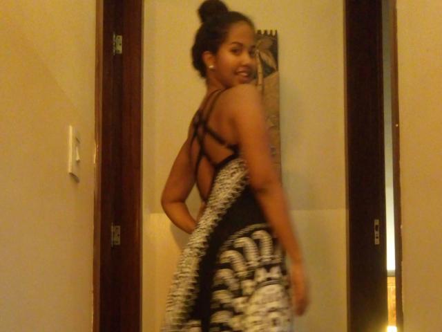LunaMelynda-hot with dark hair in a bun, smiling, wearing a black strappy top and patterned black/white pants, showing her ass, standing in a doorway.
