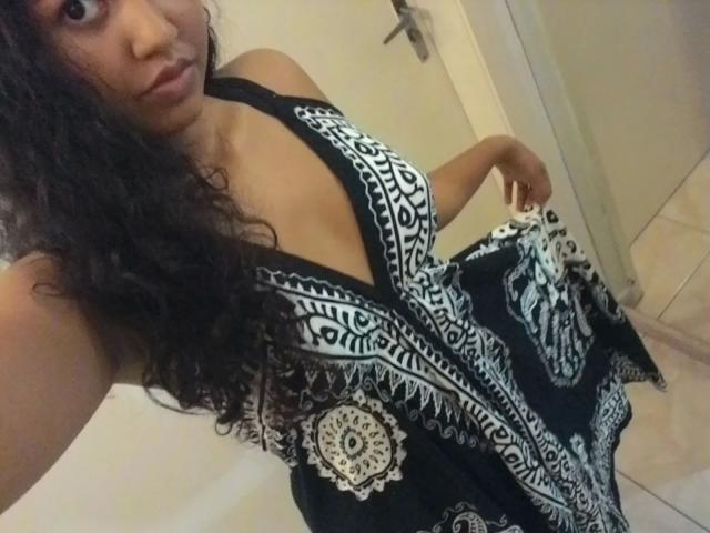 LunaMelynda-hot with curly black hair, wearing a black and white patterned dress with a deep V-neck, showing her tits, holding the dress slightly up.