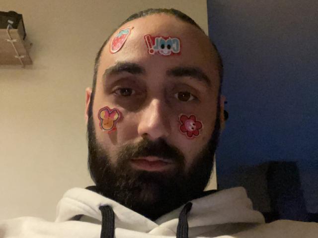 MaxoUo ha la testa rasata, una barba scura e indossa una felpa bianca. Il suo viso è ricoperto di adesivi colorati, tra cui una "I" blu e fiori rossi..