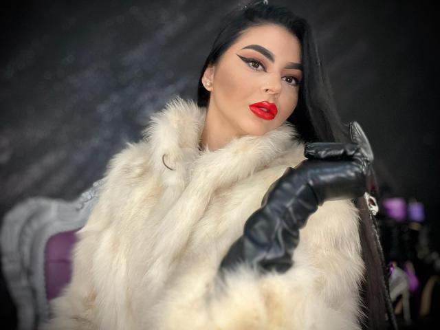 Mistress Edith Fuchs, con lunghi capelli neri, rossetto rosso e un audace trucco per gli occhi, indossa un cappotto di pelliccia bianca e guanti di pelle nera, appare seducente e sensuale..