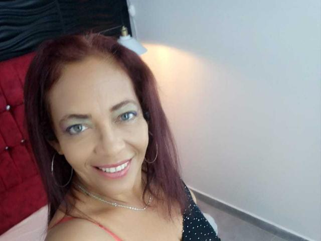 ZoeGreyX with red hair, blue eyes, smiling, wearing a black polka-dot top, red strap, hoop earrings, and a silver necklace, in a bright room.