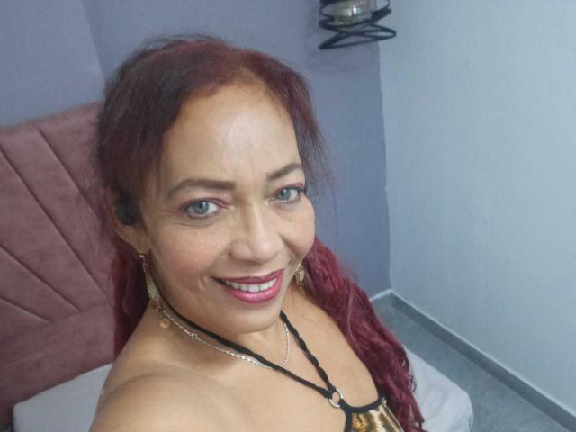 ZoeGreyX has red hair, green eyes, and is smiling at the camera with pink lipstick. She's wearing gold earrings and a black necklace with a gold clasp.