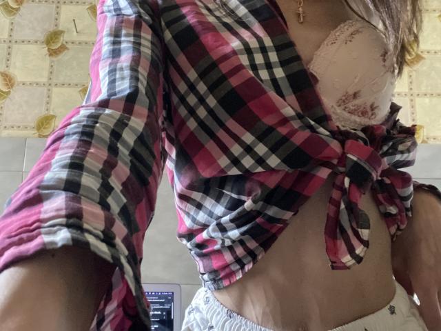 Kabblasha indossa una camicia a quadri rosa e nera, annodata davanti, che lascia intravedere dei seni bianchi ricoperti di pizzo, con una collana a croce e pantaloni da pigiama grigi..