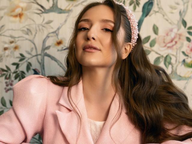 BiancaHotLove with long brown hair, pink pearl headband, pink blazer, and subtle makeup, looking slightly up with a soft smile. Boobs not visible. Background floral pattern.