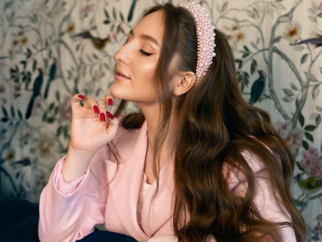 BiancaHotLove with long brown hair, pink headband, pink robe, red nails, and a cute smile, holding her chin. She's got a sexy, cute look.
