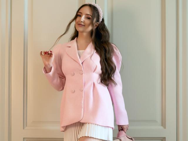 BiancaHotLove with long brown hair, pink hairband, pink jacket, white pleated skirt, pink shoes, standing, smiling, holding hair, looking cute, soft pink colors, elegant, stylish.