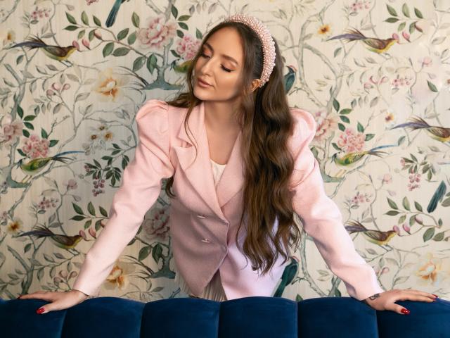 BiancaHotLove with long brown hair, pink headband, pink blazer, white shirt, leaning on blue velvet couch, eyes closed, hands on couch, nails painted red, soft smile, pretty.