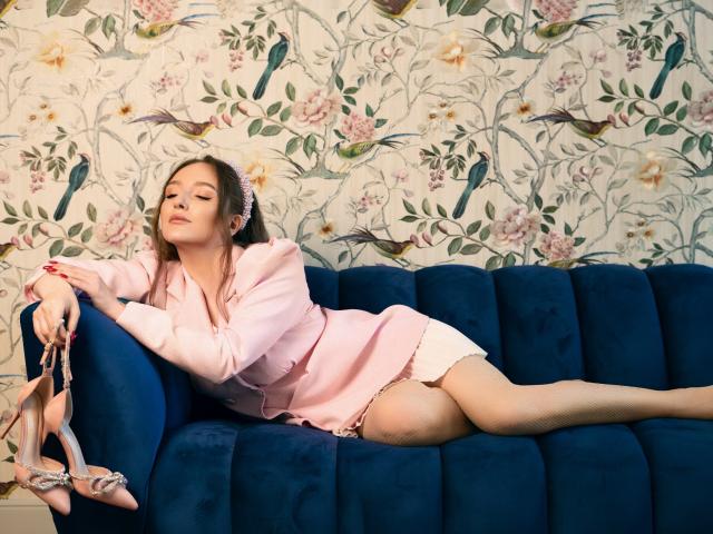 BiancaHotLove, with tits covered, lays on a blue couch in a pink dress, holding pink high heels, legs spread, wearing white fishnet stockings, floral wallpaper in background.