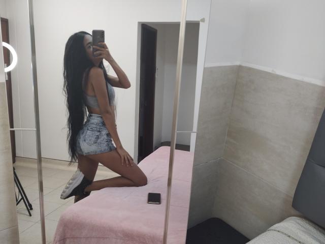 MarySmiith with long black hair, wearing a gray bra and denim shorts, kneeling on a pink-covered bed, taking a mirror selfie, showing her ass and white sneakers.