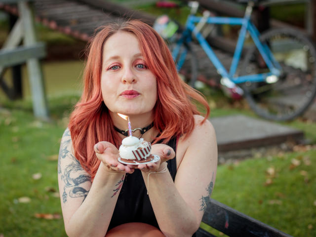 VioletaMora with red hair, tattoos on arms, black sleeveless top, pouty lips, holding white cupcake with candle, outdoors, blue bike in background.