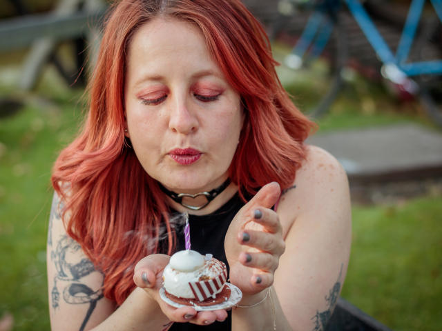 VioletaMora with red hair, pink eyeshadow, black choker, tattoos, and gray nail polish, kissing a white-frosted cupcake with pink candle, holding it with her right hand.