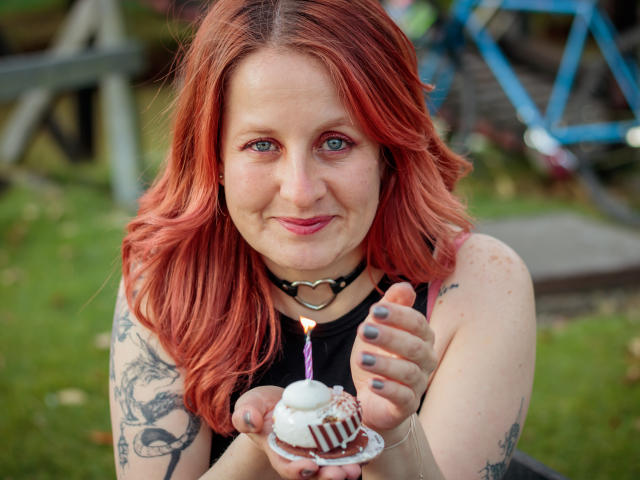 VioletaMora with red hair, blue eyes, and tattoos, holding a cupcake with a candle, wearing a black top and heart choker, smiling, outdoors, with painted nails.