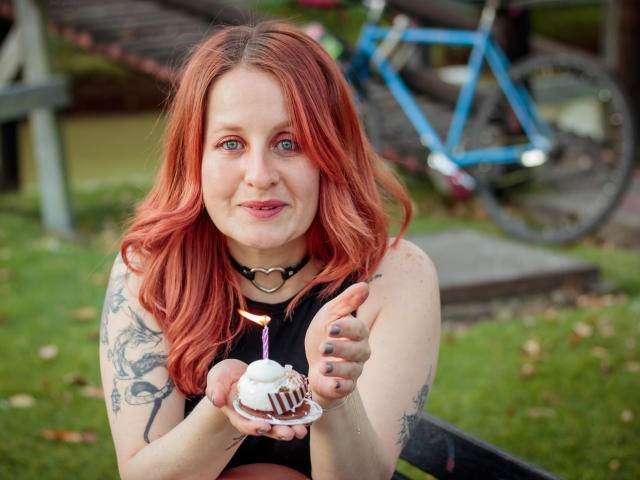 VioletaMora with red hair, holding a small white chocolate cake with a pink candle, wearing a black choker and sleeveless top, tattoo on right arm, smiling. Blue bike in background.