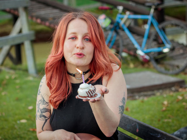 VioletaMora with red hair, black sleeveless top, and tattoos, holding a cupcake with a candle, pink lipstick, and a black choker, sitting on a bench, outside.