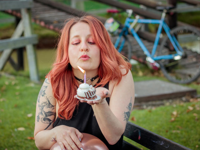 VioletaMora with red hair, tattoos on arms, black sleeveless dress, blowing out candle on cupcake, blue bicycle in background, sitting on black bench, holding cupcake in right hand.