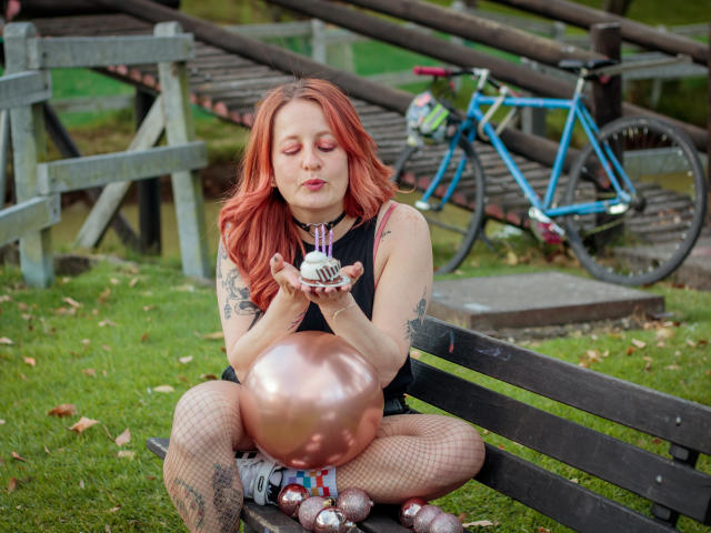VioletaMora with red hair, eyes closed, peeks over a gray barrier, blue bicycle with green and pink accessories in the background.