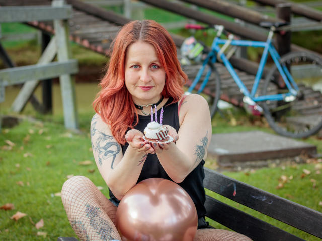 VioletaMora with red hair, tattoos on arms, fishnet stockings, black sleeveless top, holding cake with purple candles, sitting on bench with metallic balloon, blue bike in background.