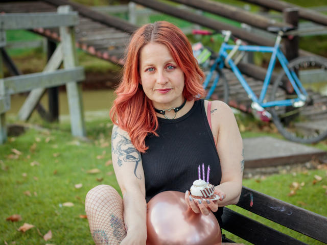 VioletaMora with red hair peeks over a gray barrier, her blue eyes and slight smile visible, with a blue bike in the background.
