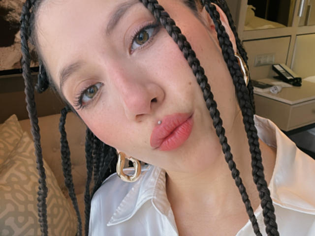 IzzysMisss has braided black hair, a nose piercing, and pink lipstick. She's wearing a white shirt with gold earrings, looking at the camera with pouty lips.