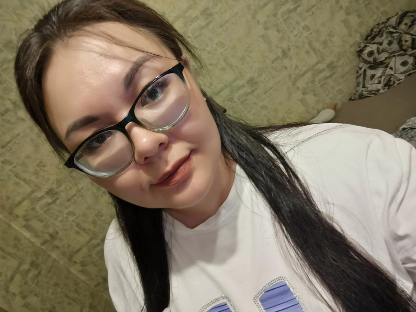 Denise, con cabello negro largo, gafas y una camisa blanca, sonriendo ligeramente, mostrando una piel clara y un rostro lindo, con sus senos parcialmente visibles..