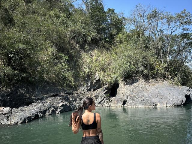 ThaliaAres, avec un tatouage sur le dos, un débardeur de sport noir et un pantalon noir, se tient dans une rivière verte, face à une berge rocheuse et boisée, tenant un sac marron dans sa main droite..