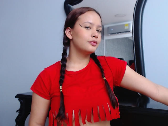 Lauren Martins, con i capelli castano intrecciati, un top corto rosso con frange e un anello al naso nero, è in piedi di fronte a uno specchio, guardando leggermente di lato con un'espressione sicura..