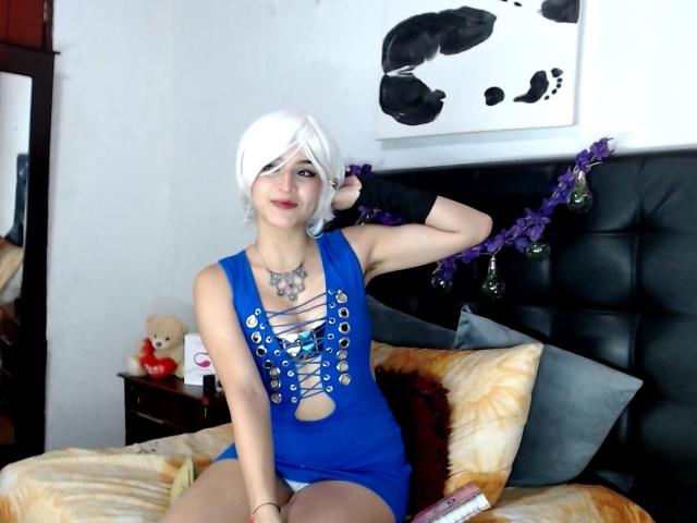 CollinsCharlotte with white bob, blue dress, lace-up cleavage, black gloves, sitting on bed, hand in hair, purple garland, teddy bear, and white wall art, showing tits.