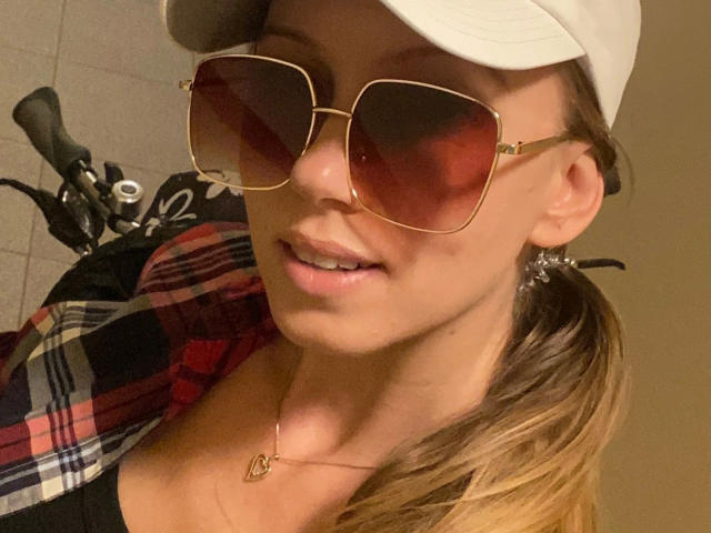 Julie Mayer, mit Sonnenbrille, weißem Hut, kariertem Hemd und Halskette, zeigt ihre vollen Lippen und ihr dezentes Dekolleté, im Hintergrund ist ein Fahrrad zu sehen..