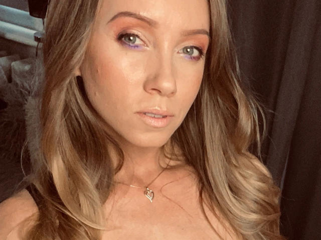 JulieMayer-hot heeft blond haar, groene ogen, paarse oogschaduw en een hartvormige ketting. Haar borsten zijn licht zichtbaar, en ze kijkt recht in de camera met een neutrale uitdrukking..