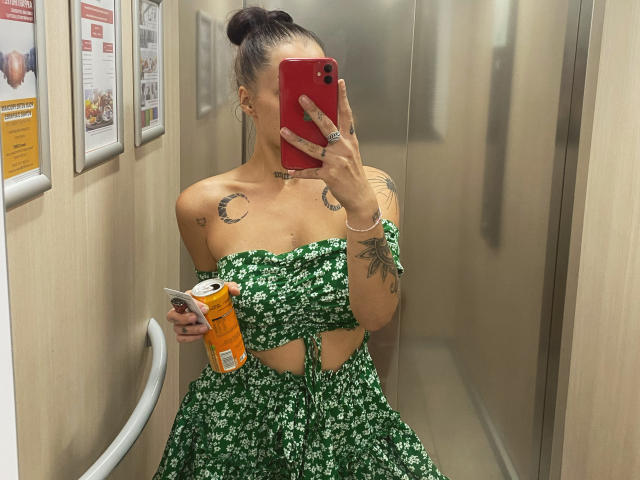 Sacha69Hot with tattoos, green floral dress, off-shoulder, holding orange can, red phone, mirror selfie, elevator, brown bun, visible chest, ring on left hand.