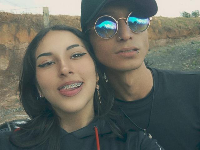 SrgreyAndJulieta with dark hair, black hoodie, and earrings, tongue out with metal teeth, standing next to SrgreyAndJulieta in black cap, reflective sunglasses, black shirt, and nose ring.