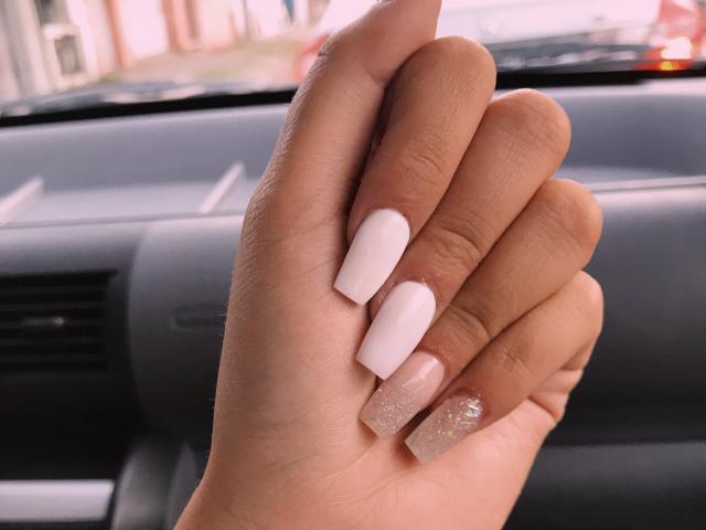 A mão de AlexaLip, com unhas longas e rosa pálido, uma delas com brilho, dentro de um carro. Sua pele é bronzeada, os dedos levemente curvados. O foco está em suas unhas bem cuidadas..