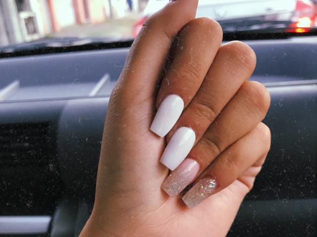 AlexaLip's hand with white and pink glitter nails, slightly curled fingers, dark skin, and a small visible cuticle, in a car with a window in the background.