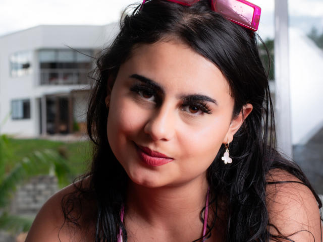 LeilaEvanns has dark, wavy hair, pink lipstick, and is wearing pink sunglasses on her head. She has small, white star earrings and a pink halter top.