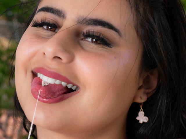 LeilaEvanns' face, tongue out, drooling a thick string of saliva, dark hair, pink lipstick, black eyeliner, pink flower earrings, light brown skin, and dark eyebrows.