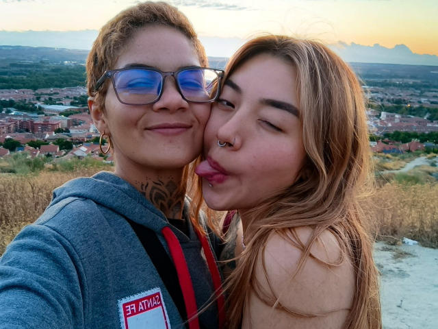 SeleneAna with short curly hair, blue glasses, and a grey hoodie, standing next to SeleneAna with long blonde hair, nose ring, and tongue out, in a sunset selfie.