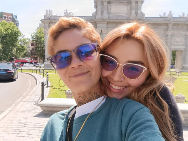 SeleneAna with short orange hair and blue sunglasses, tattoo on neck, wearing teal sweater, stands beside SeleneAna with long blonde hair, white sunglasses, nose ring, smiling.