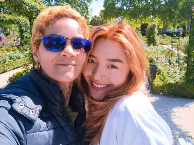 SeleneAna with short orange hair, blue sunglasses, black puffy jacket, and tattoos, and SeleneAna with long red hair, nose piercing, and white shirt, smiling in a sunny garden.