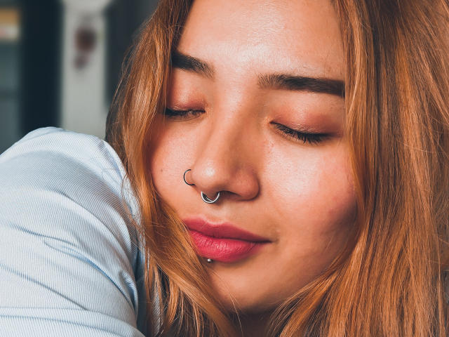 SeleneAna with closed eyes, wearing a nose ring and lip piercing, has pink lipstick and light brown hair. She's in a light blue shirt, looking relaxed.