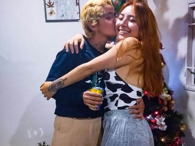 SeleneAna with red hair, small tits, wearing a black-and-white cowprint top, hugs a blonde man with glasses, tattoos, and a dark shirt, kissing her, holding a can.