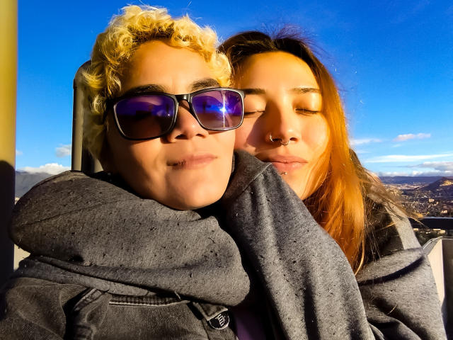 SeleneAna with curly blonde hair and purple sunglasses, arms around a brunette SeleneAna with nose ring, both wearing grey hoodies, eyes closed, sunlight, clear blue sky, happy.