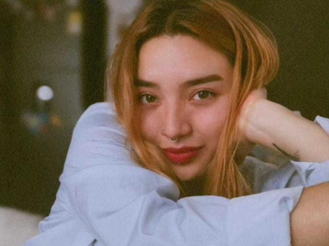 SeleneAna with light brown hair, nose ring, and blue shirt, leaning on her hand, smiling softly, brown eyes, subtle makeup, relaxed and cute, medium shot.