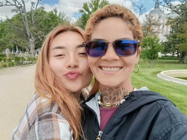 SeleneAna with pink hair and nose ring, pouty lips, wearing plaid shirt, next to SeleneAna with curly brown hair, tattoos, sunglasses, black jacket, and gold hoop earrings.