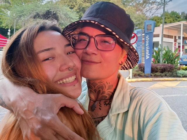 SeleneAna with long brown hair and nose ring, smiling, being hugged by a tattooed person with glasses, black bucket hat, and green-striped shirt. SeleneAna's hand rests on her shoulder.
