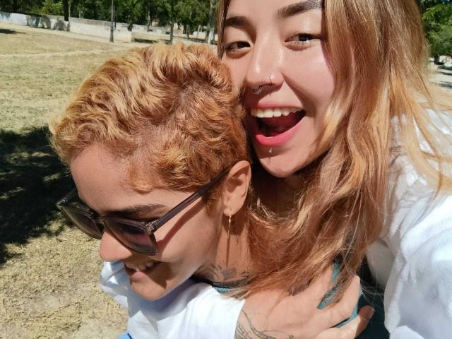 SeleneAna with blonde curly hair and nose ring, smiling, leans over a guy with glasses, white shirt, and short curly red hair, both outside in sunny park.