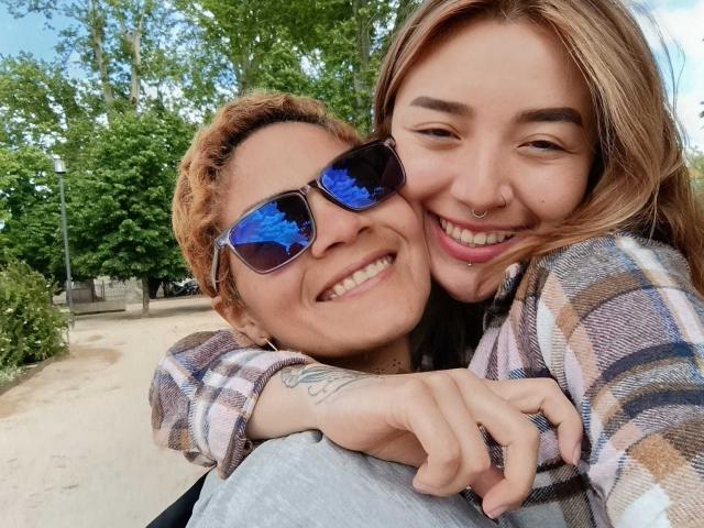 SeleneAna with short, curly brown hair and sunglasses, smiling with her nose pierced, hugging SeleneAna with blonde hair and a plaid shirt, showing a tattoo on her arm.